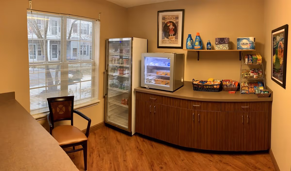 Small kitchenette/snack area with a refrigerated display case, countertop snacks, a chair, and a window overlooking nearby buildings.