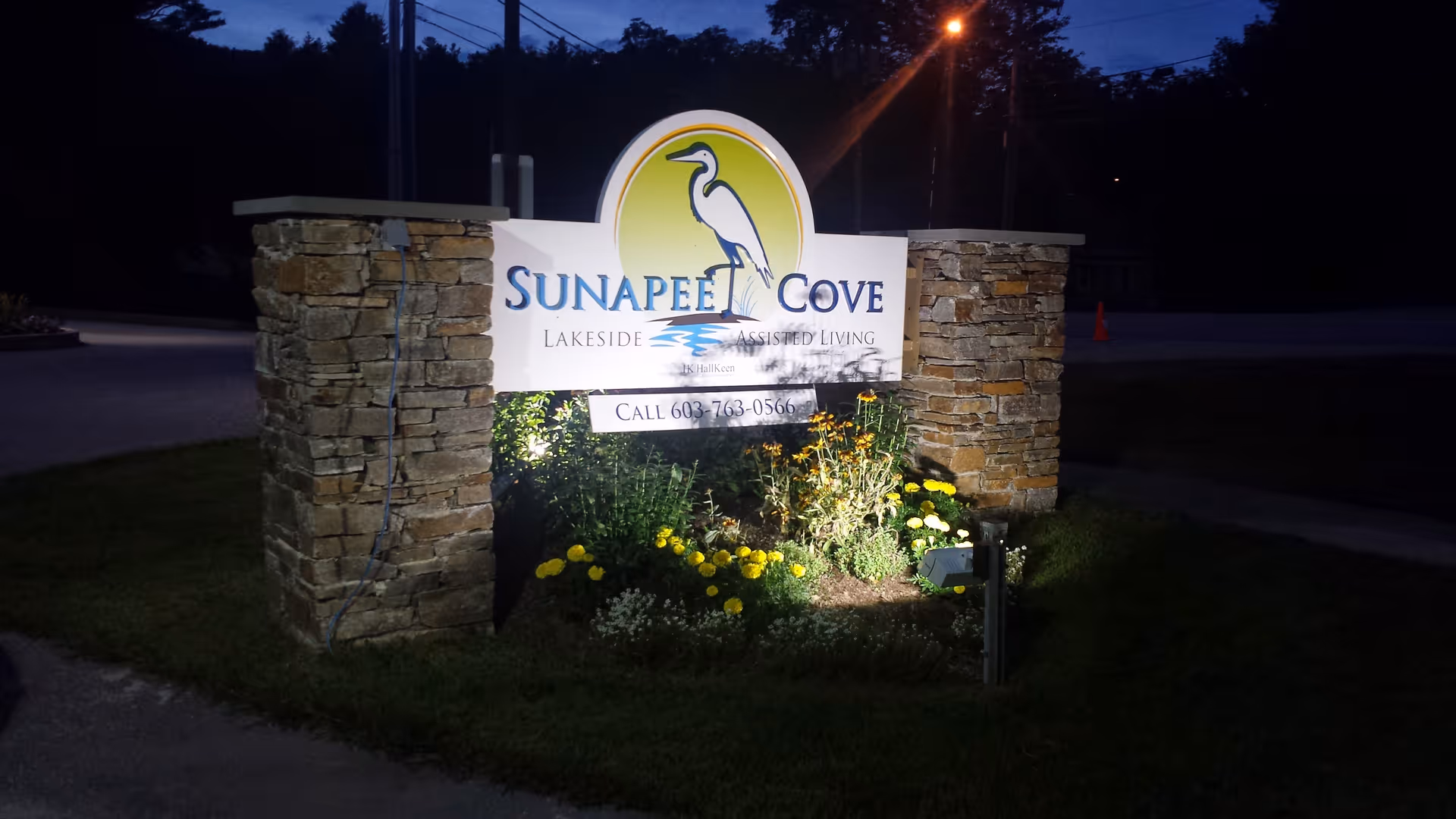 Illuminated stone entrance sign for Sunapee Cove Assisted Living at night, flanked by stone pillars and flowerbeds.