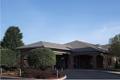 Single-story brick building with a covered porte-cochère entrance, trimmed shrubs, and a circular driveway under a clear blue sky.