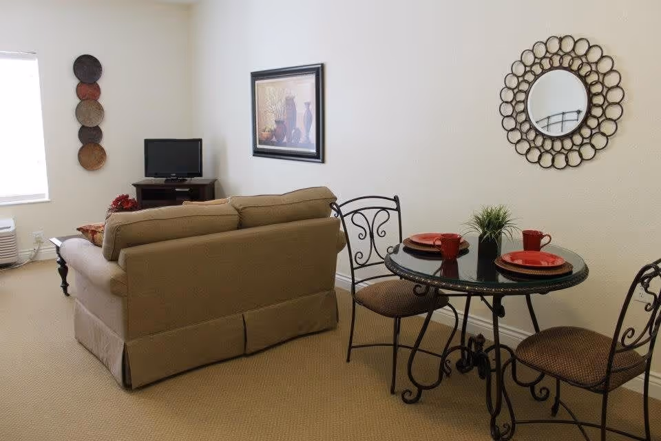A cozy living area featuring a beige sofa facing a small TV on a wooden stand. To the right, there is a round glass-top dining table with two wrought iron chairs, set with red plates and mugs. The wall behind the table has a decorative circular mirror, and the opposite wall has a framed artwork and a vertical arrangement of decorative plates. The room has beige carpeting and white walls.