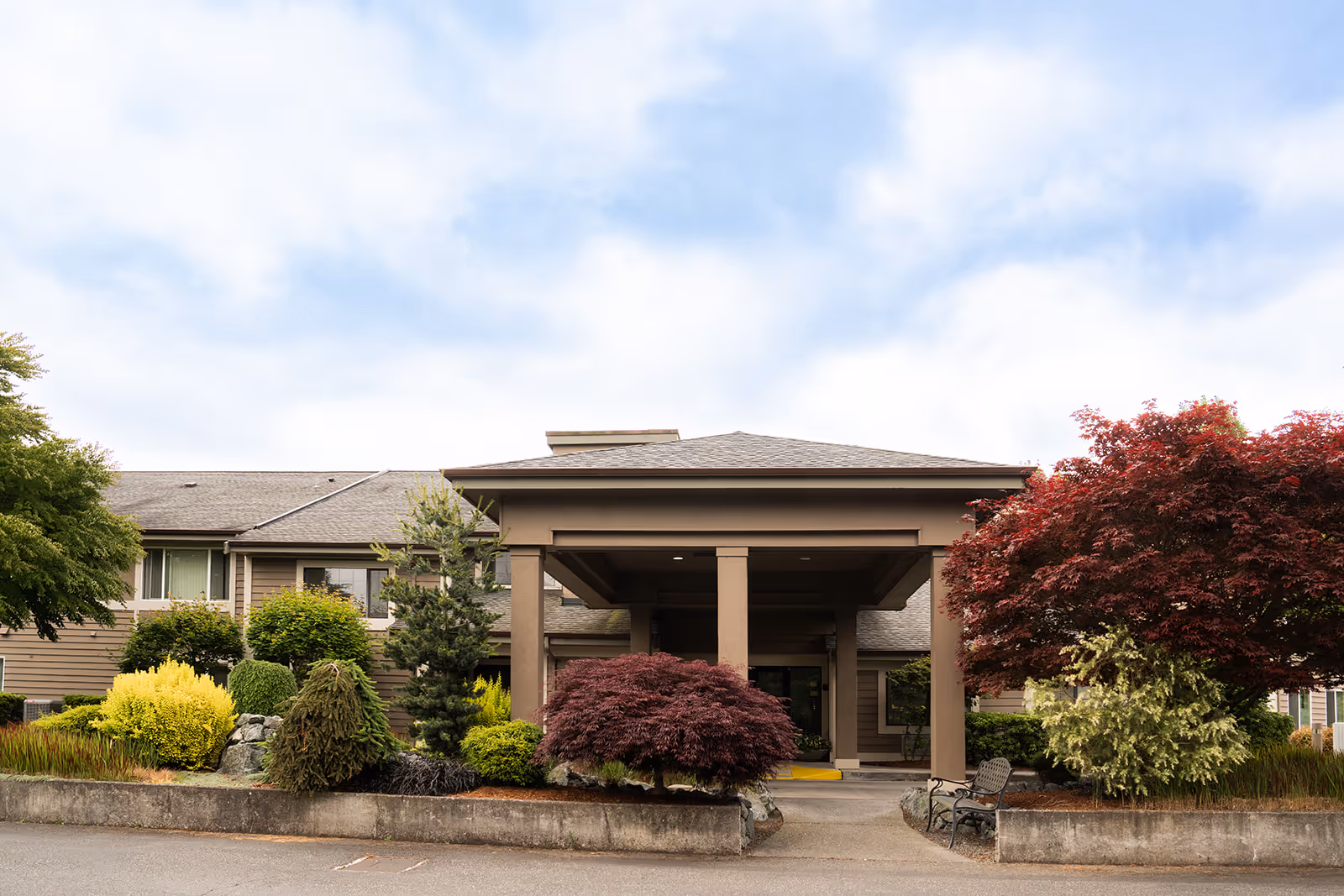 Front exterior view of Village Concepts of Sedro-Woolley - Country Meadow Village building with a covered entrance, surrounded by well-maintained landscaping including various shrubs and trees under a partly cloudy sky.