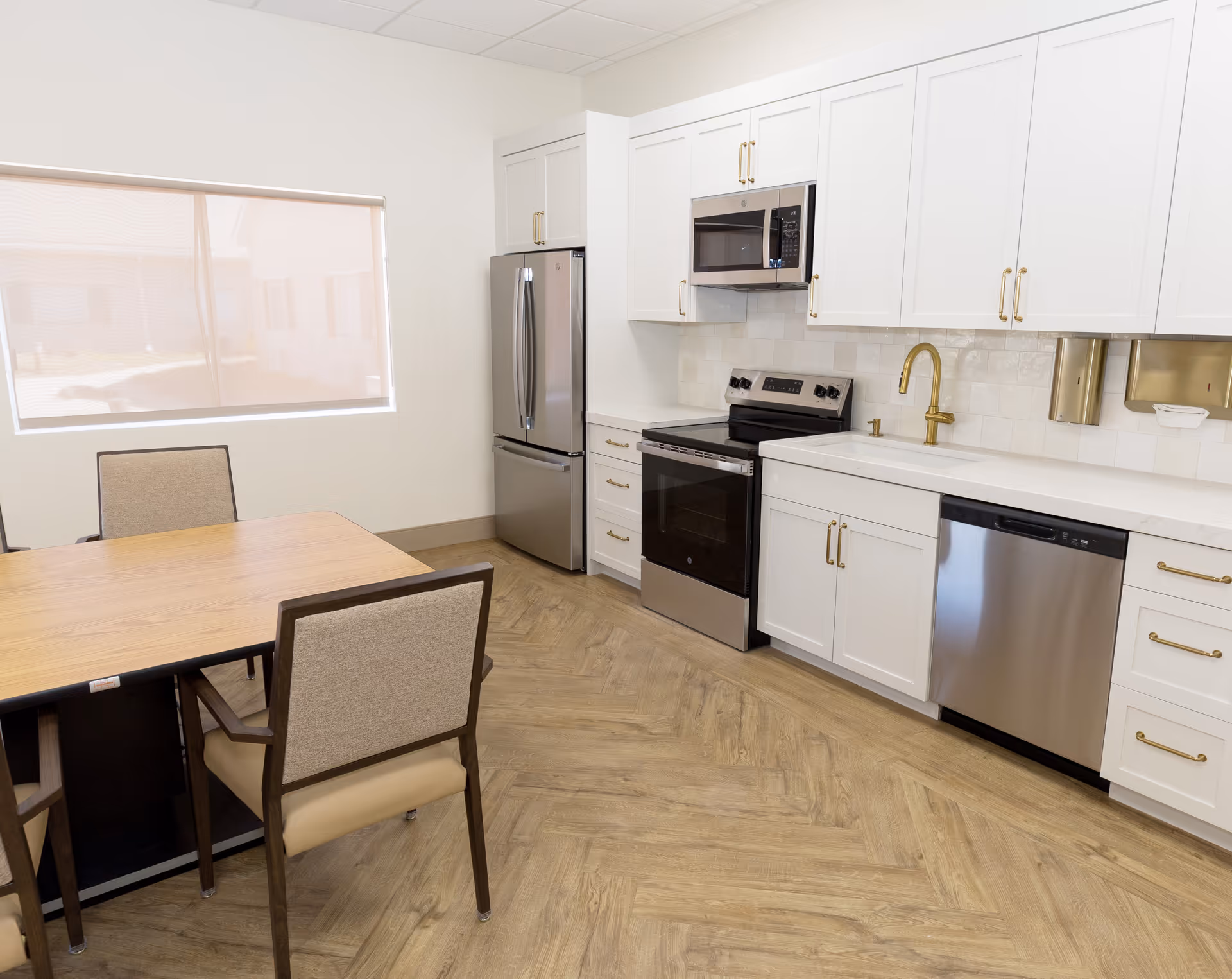 A modern kitchen with white cabinets, stainless steel appliances including a refrigerator, oven, microwave, and dishwasher. There is a wooden dining table with four cushioned chairs, and a window with a beige roller shade letting in natural light.