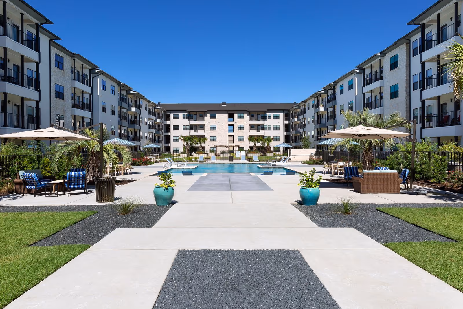 Outdoor courtyard area of a senior living facility with a swimming pool in the center, surrounded by lounge chairs, umbrellas, and seating areas. The courtyard is flanked by multi-story residential buildings under a clear blue sky.