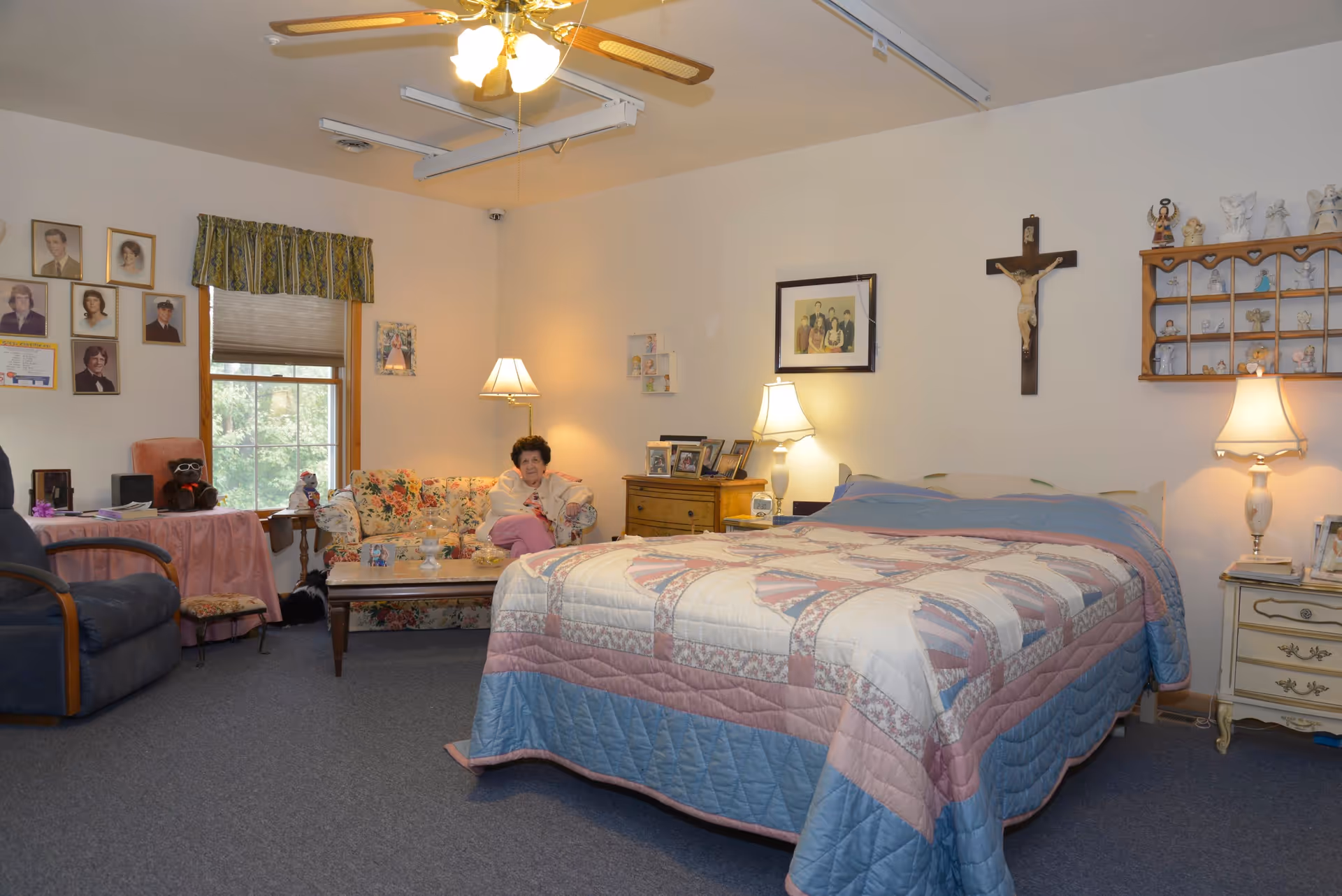 A cozy bedroom in an assisted living facility featuring a large bed with a quilted bedspread, two bedside tables with lamps, a floral couch with an elderly woman sitting on it, a recliner chair, a coffee table, and various framed photos and religious decorations on the walls.