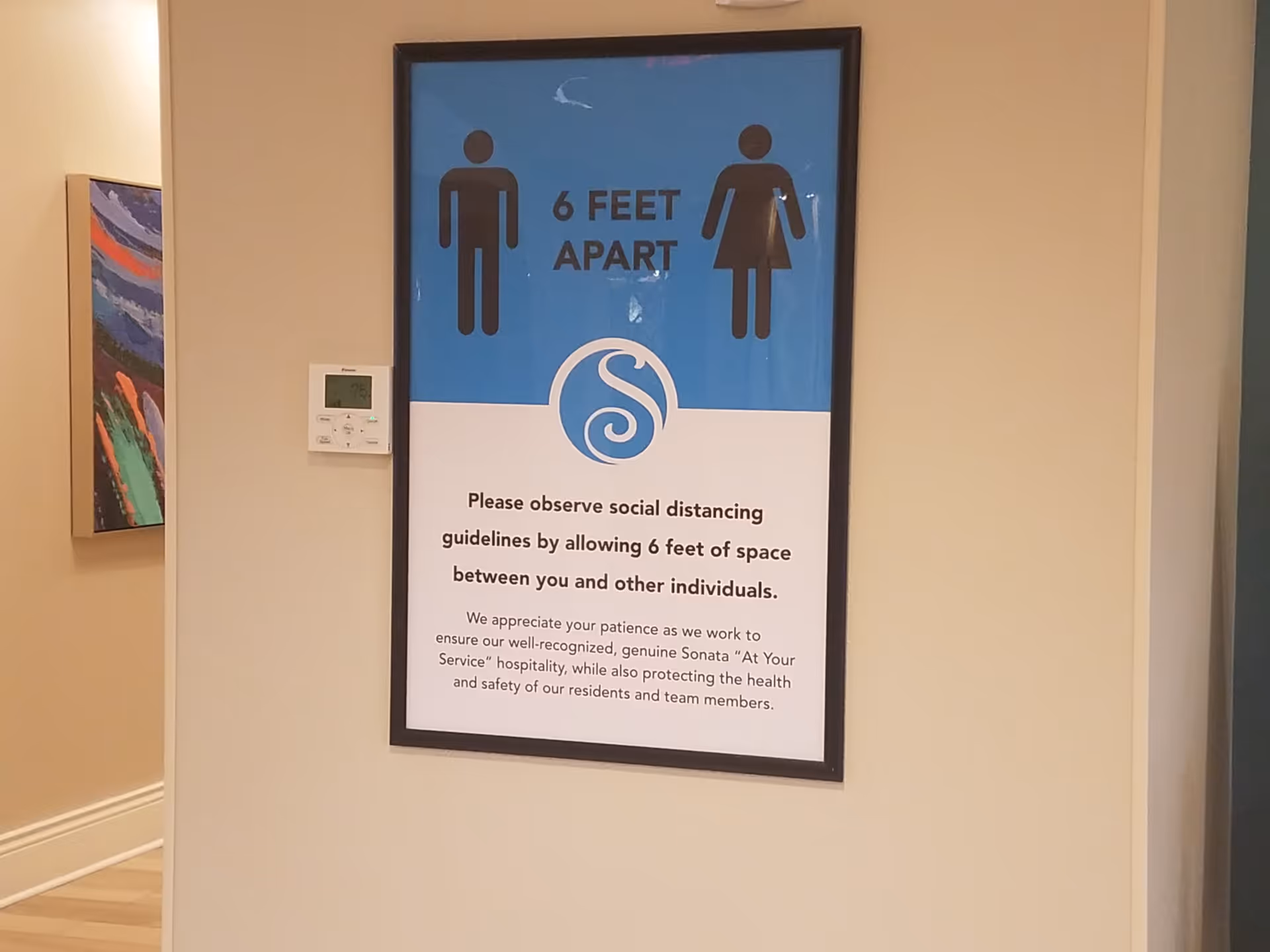 A framed sign on a beige wall instructing to maintain 6 feet apart for social distancing. The sign includes icons of a man and a woman, the text '6 FEET APART', and a message requesting to observe social distancing guidelines by allowing 6 feet of space between individuals. There is a thermostat on the wall to the left of the sign and a colorful painting partially visible in the adjacent hallway.