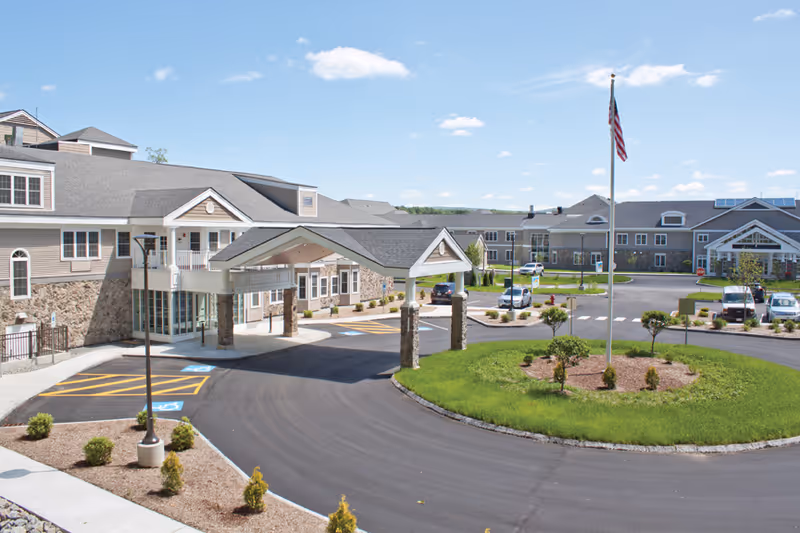 Exterior view of The Prospect-Woodward Health Center at Hillside Village showing a large building with a covered entrance, a circular driveway with a flagpole in the center, several parked cars, and a clear blue sky.