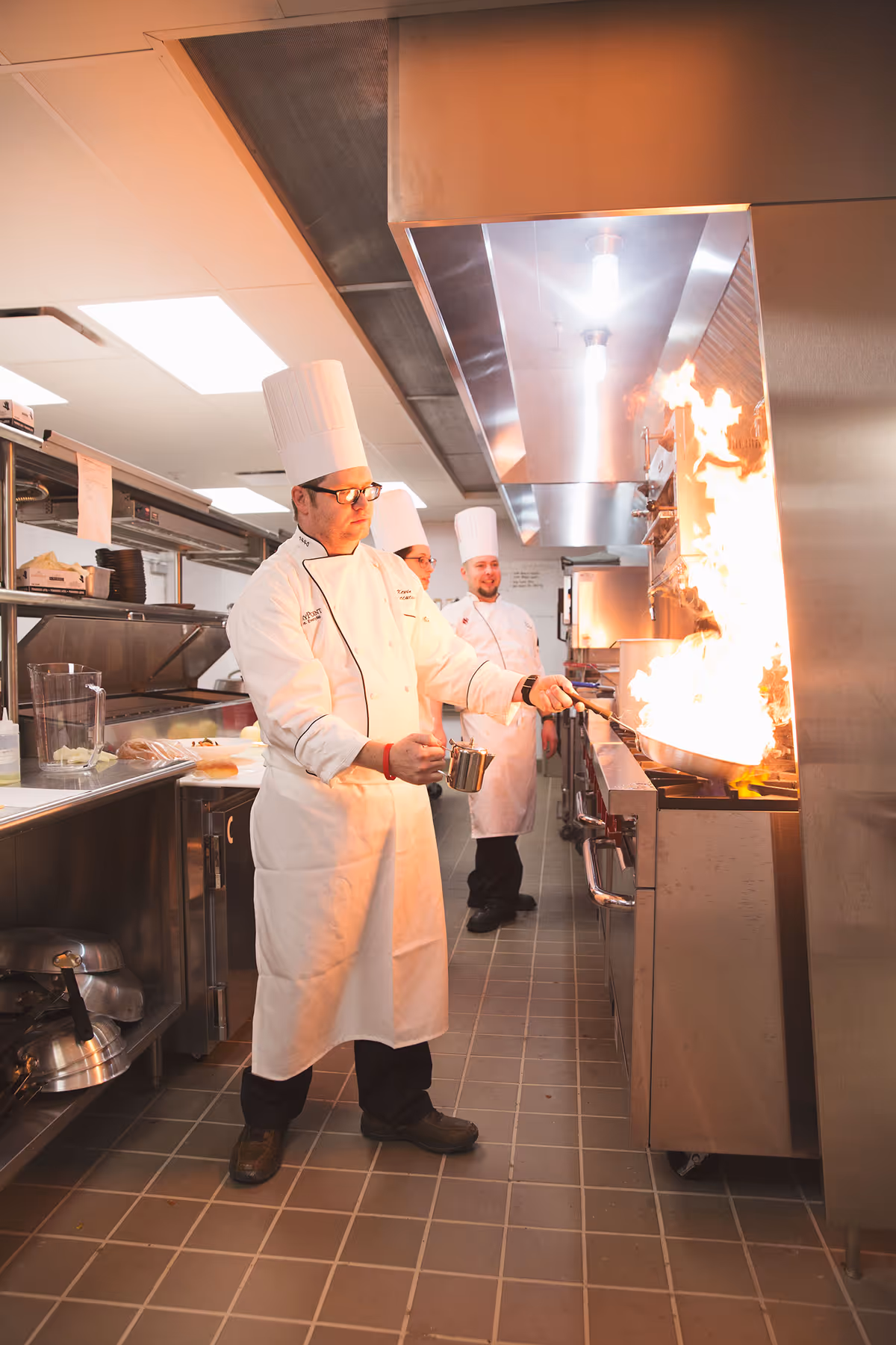 Three chefs in white uniforms and tall white chef hats working in a commercial kitchen. One chef is flambéing food in a pan, creating a large flame. The kitchen has stainless steel appliances and tiled floors.
