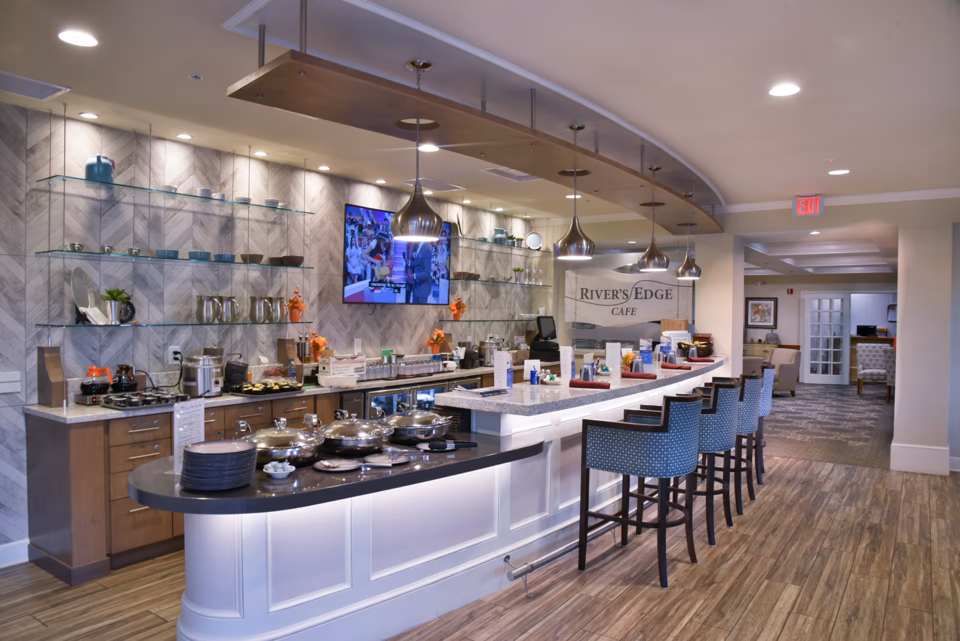 Interior view of a modern cafe area with a curved counter and several high chairs. The counter has various food warmers and plates, with a backdrop of shelves holding cups and decorative items. A TV is mounted on the wall above the counter, and pendant lights hang from the ceiling. The sign on the wall reads 'Rivers Edge Cafe'.