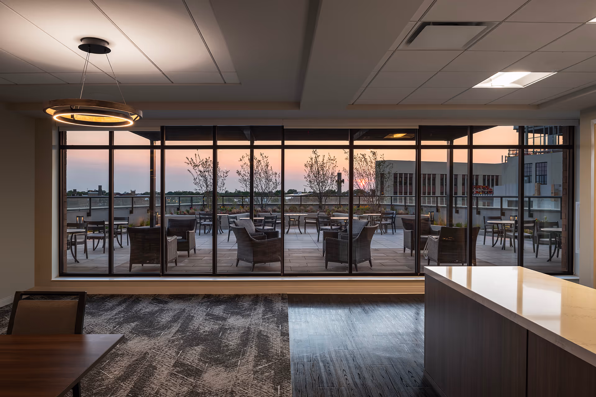 Interior view of a modern room with large floor-to-ceiling windows looking out onto a spacious outdoor patio area with multiple tables and chairs. The patio has potted plants and trees, and the sun is setting in the background. Inside, there is a wooden table with a chair and a countertop with a smooth surface. A circular ceiling light fixture is also visible.