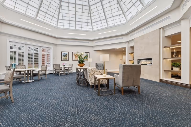 A spacious and well-lit common area with a large skylight ceiling, blue patterned carpet, multiple seating arrangements including armchairs and small tables, a modern fireplace built into the wall, and decorative plants and framed artwork on the walls.