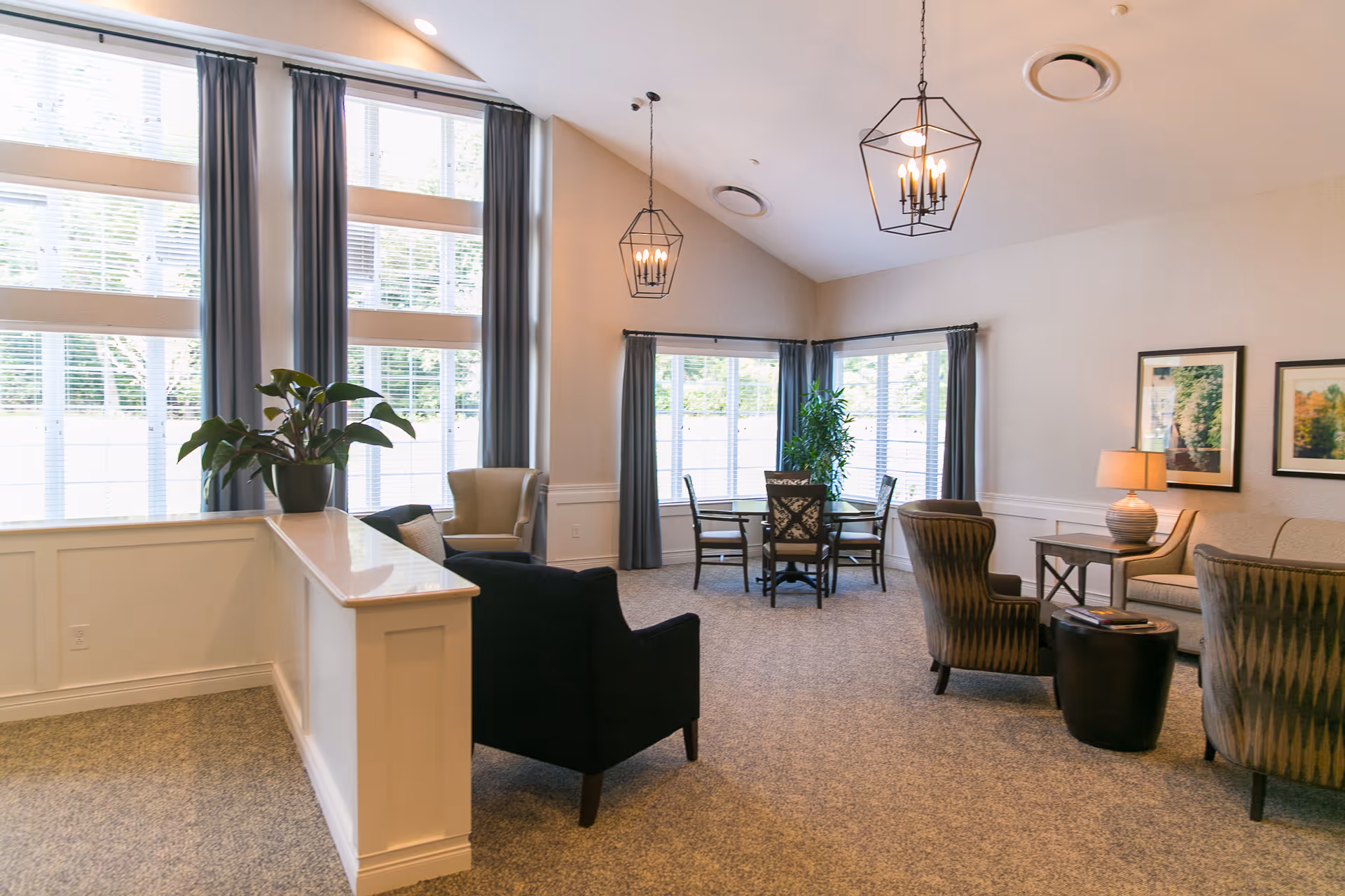A bright and spacious senior living common area with large windows covered by blue curtains, multiple seating options including armchairs and a sofa, a round table with chairs near the windows, two hanging pendant lights, framed artwork on the wall, a table lamp, and a potted plant on a white half-wall.