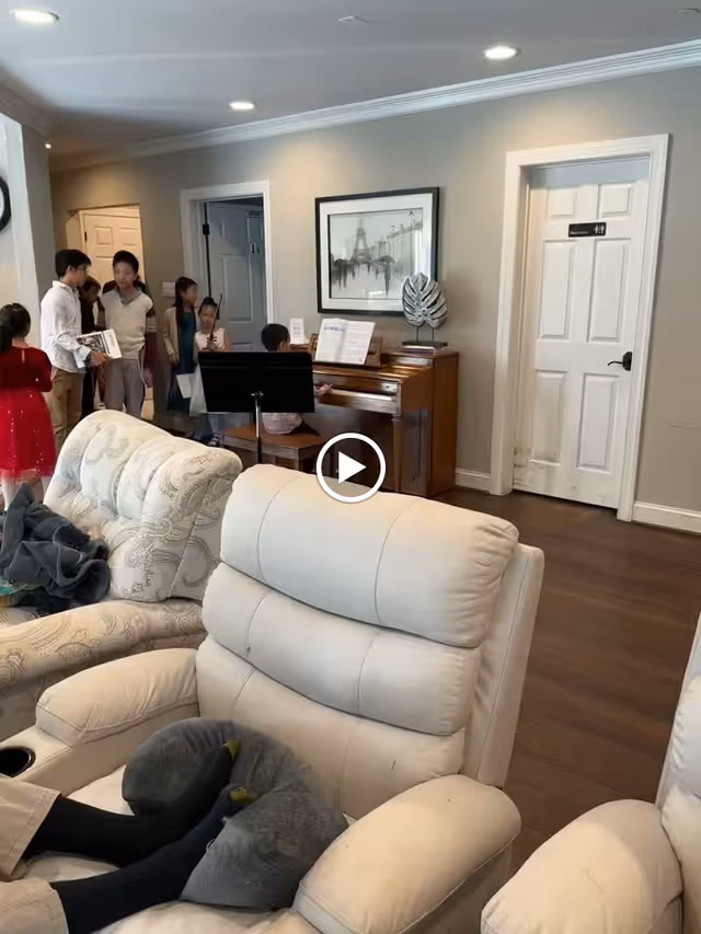 A group of children standing near a piano in a living room area. One child is playing the piano while others watch. The room has beige walls, wooden flooring, and comfortable upholstered chairs in the foreground. A framed picture hangs on the wall above the piano.