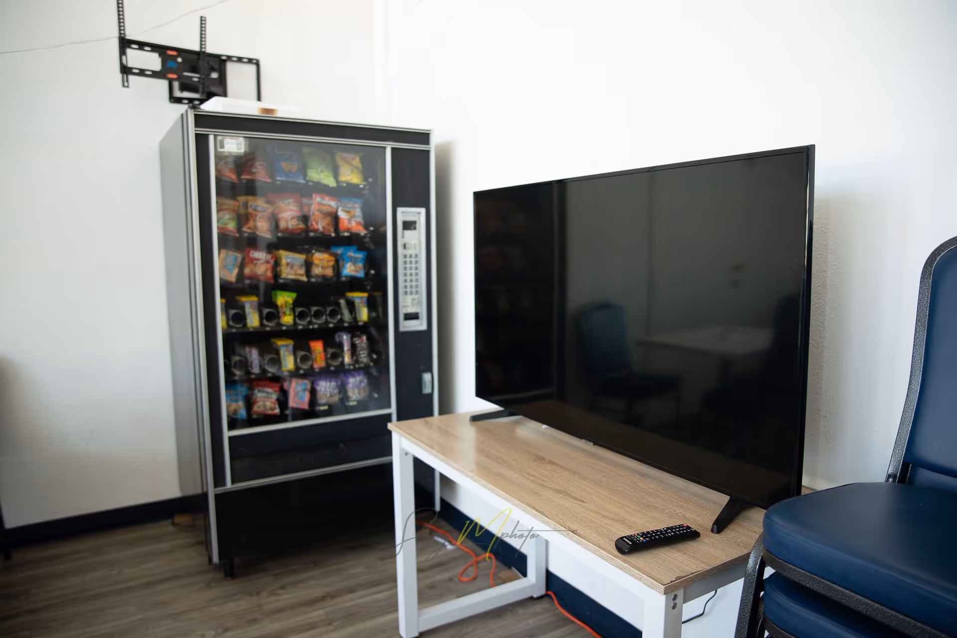 A snack vending machine filled with various chips and snacks next to a wooden table holding a large flat-screen TV and a remote control, with a blue chair beside the table in a room with light-colored walls and wood flooring.