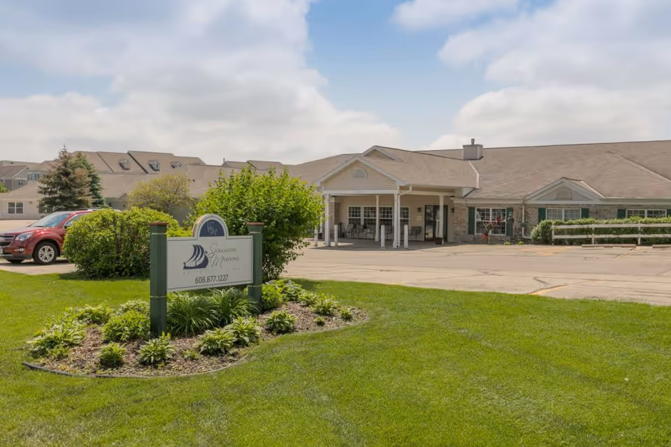 Front exterior of Stoughton Meadows assisted living showing the entrance canopy, landscaped sign, and parking area.