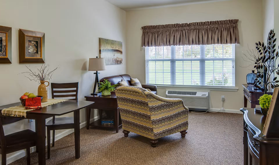 A cozy senior living room with a patterned armchair, a brown leather loveseat, a wooden side table with a lamp and plant, a small dining table with two chairs, and a large window with blinds and a valance letting in natural light.
