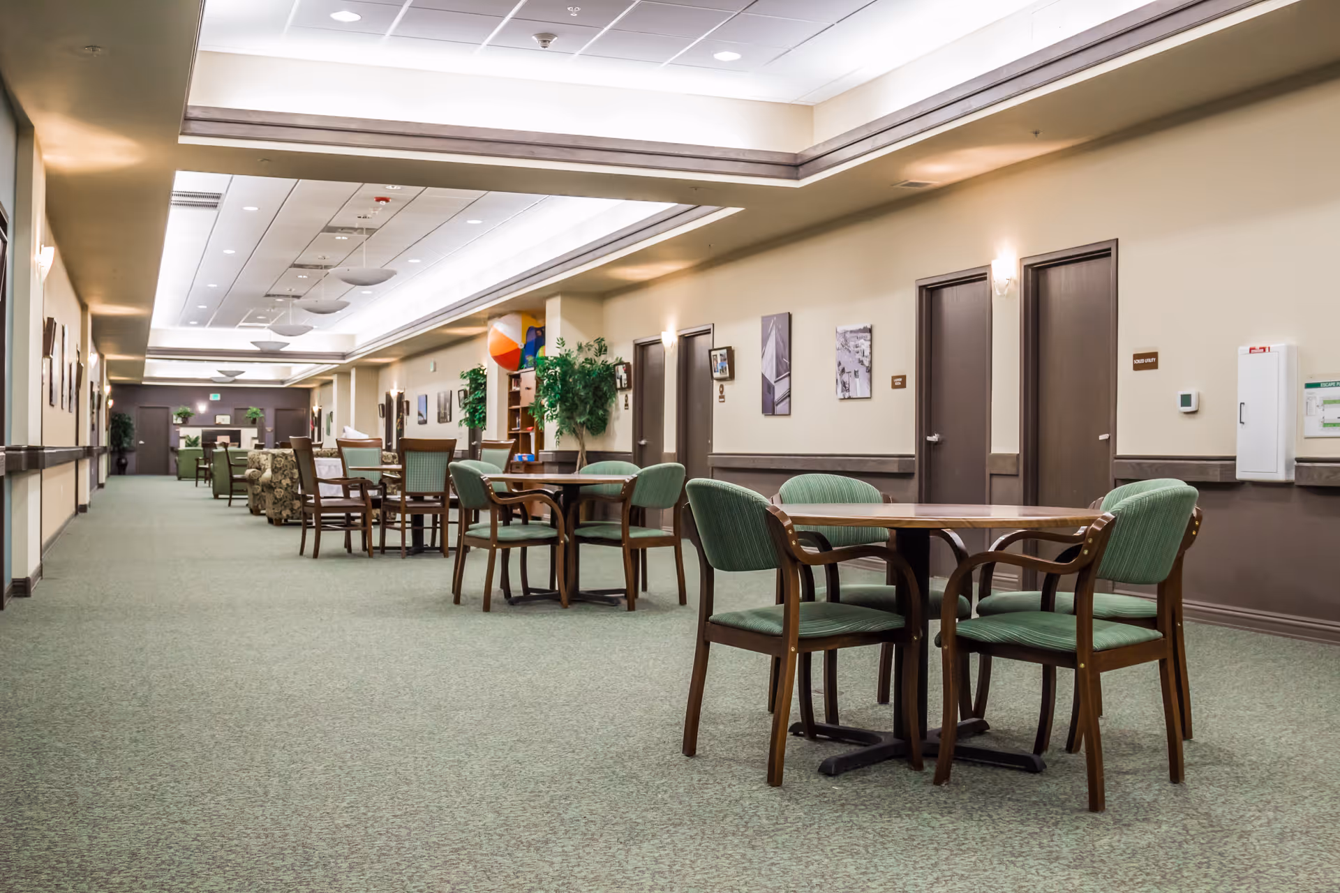 Long carpeted interior common area with round tables and green upholstered chairs arranged along a hallway under recessed ceiling lighting.