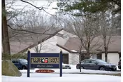 Exterior view of Apple Rehab Shelton Lakes facility with a sign in the foreground displaying the facility name. Several cars are parked near the building, and leafless trees surround the area, indicating a winter season with snow on the ground.