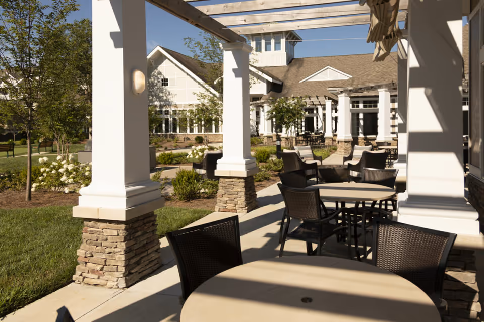 Outdoor patio area at The Heritage at Brentwood with round tables and chairs under a pergola, surrounded by landscaped gardens and white flowering bushes, with a building featuring white pillars and stone bases in the background.