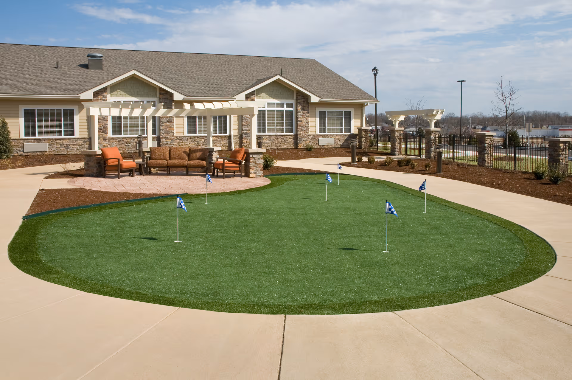 Outdoor putting green with five small flags on a well-maintained artificial turf surrounded by a concrete walkway. In the background, there is a single-story building with beige siding and stone accents, featuring large windows and a pergola with outdoor seating including a sofa and chairs. The sky is partly cloudy.