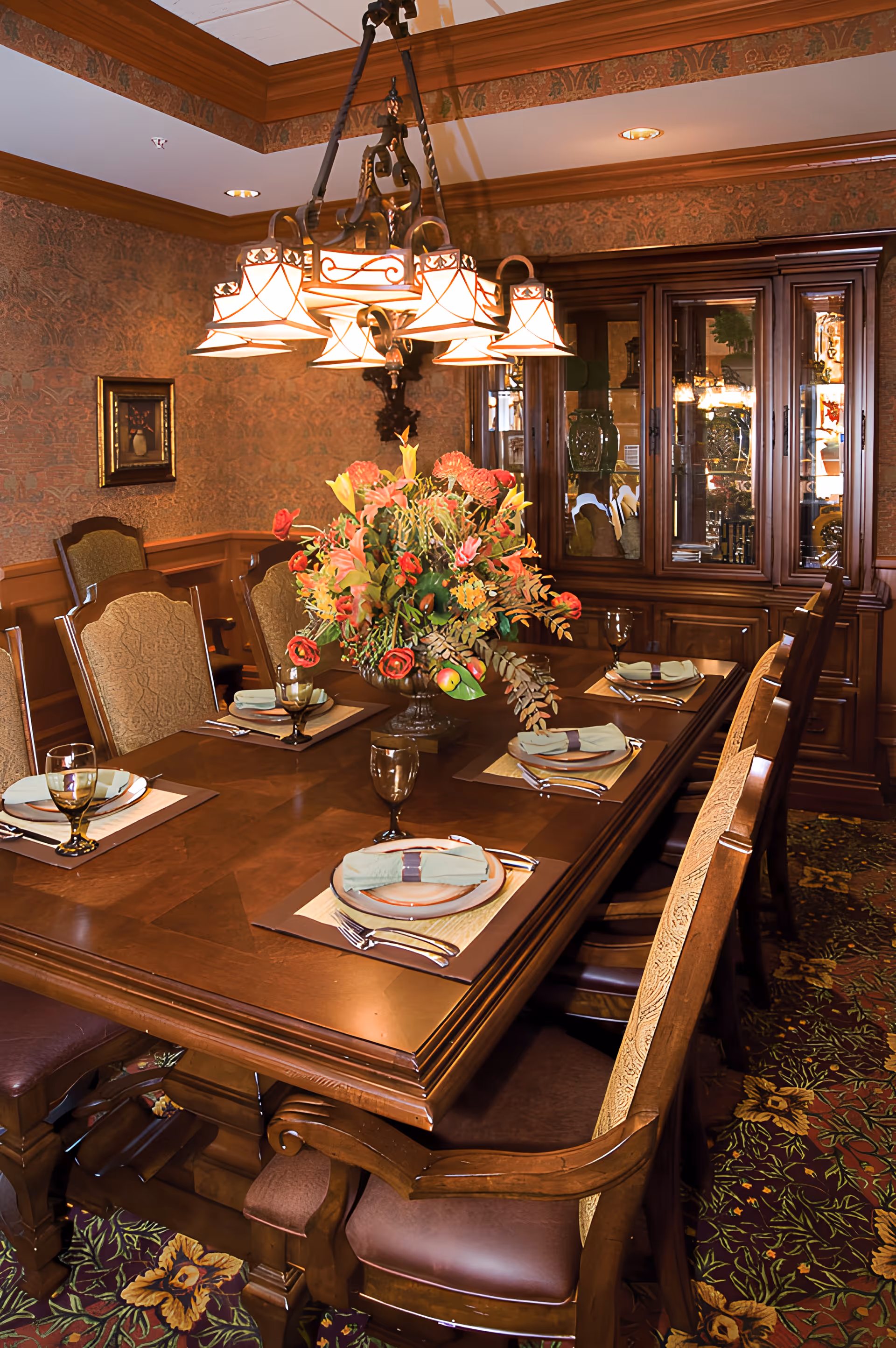 A formal dining room with a polished wooden table set with plates and glassware, a large floral centerpiece, chandelier, and a china cabinet.