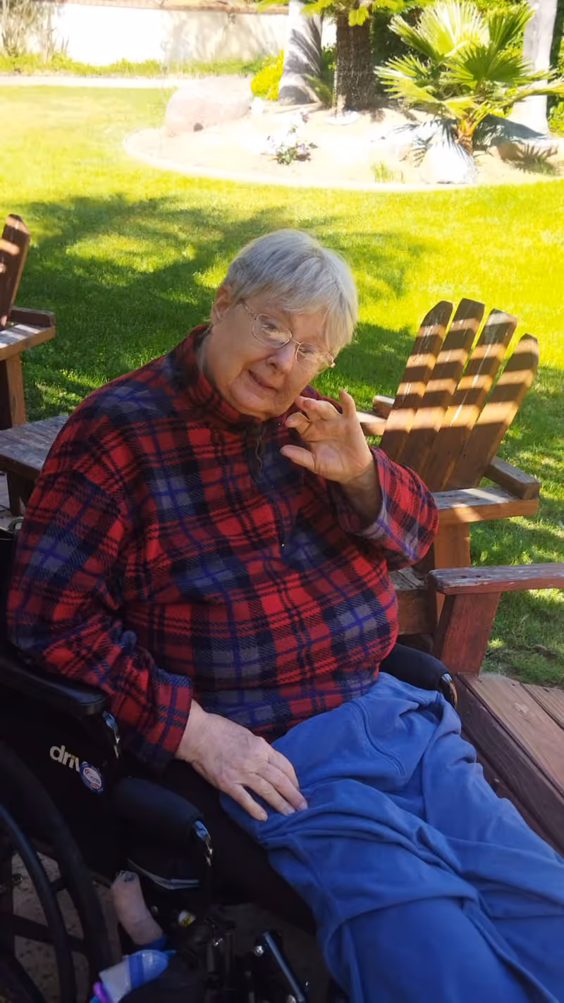 An elderly person with gray hair and glasses sitting in a wheelchair outdoors on a sunny day. They are wearing a red and blue plaid jacket and blue pants. Behind them are wooden Adirondack chairs and green grass with some plants and trees in the background.