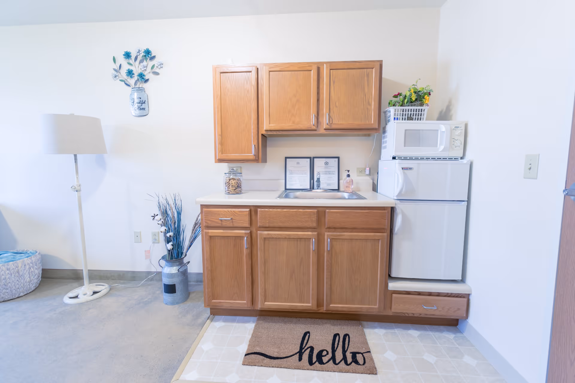 A small kitchenette area with wooden cabinets above and below a countertop with a sink. On the countertop are a jar of cookies and a soap dispenser. To the right, there is a small white refrigerator with a microwave on top and a basket with artificial flowers. A brown doormat with the word 'hello' is placed on the floor in front of the kitchenette. To the left, there is a standing lamp and a decorative vase with dried plants. The walls are white and the floor is a combination of carpet and linoleum.