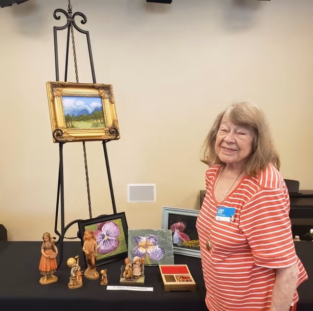 An elderly woman wearing a red and white striped shirt stands smiling next to a table displaying various small figurines and framed paintings, including floral artwork and a landscape painting on an easel.