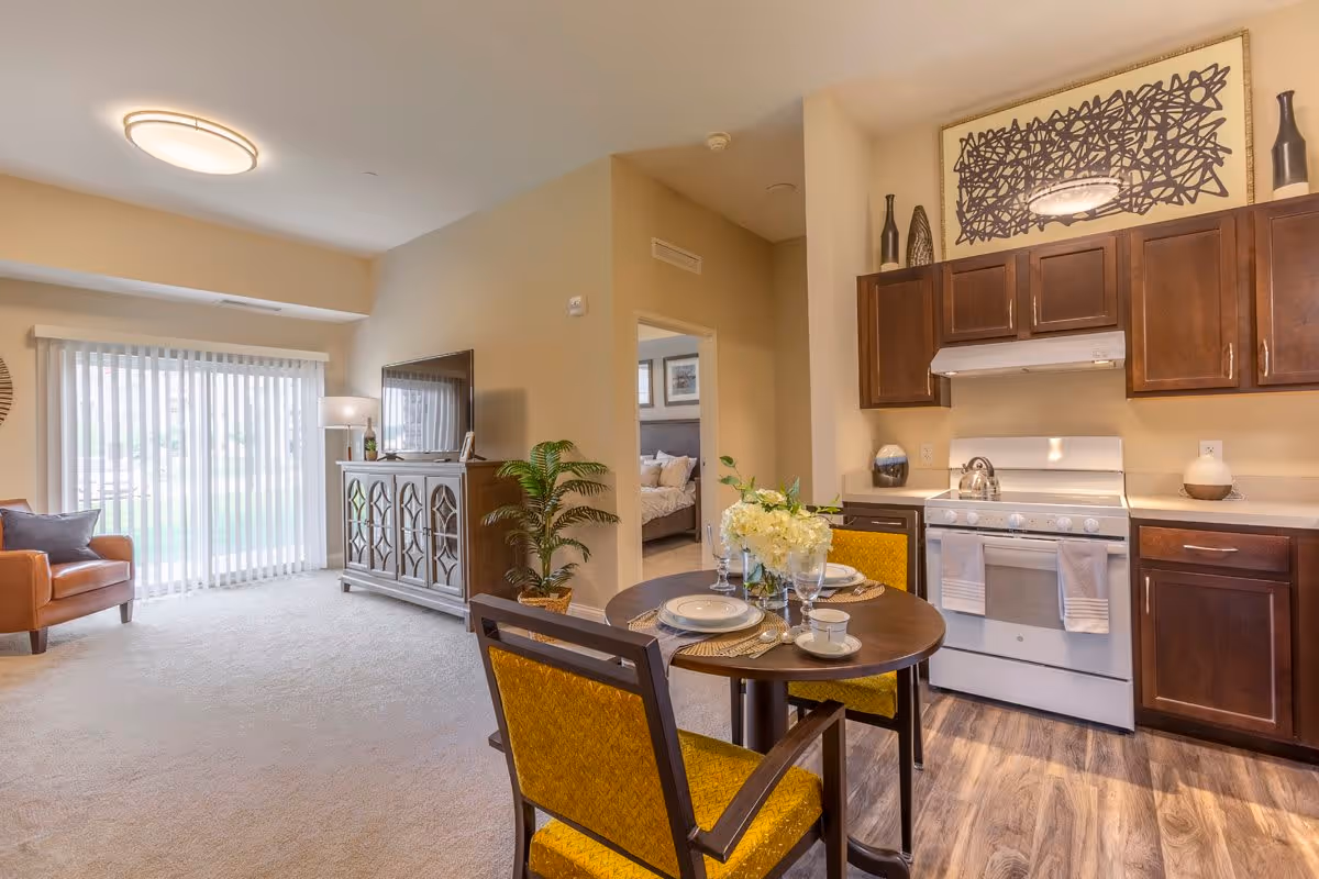 Interior view of a senior living facility apartment featuring a small dining area with a round table set for two, a kitchen with dark wood cabinets and a white stove, a living room with a brown armchair, a TV on a decorative cabinet, and a sliding glass door with vertical blinds. A bedroom is visible through an open doorway.