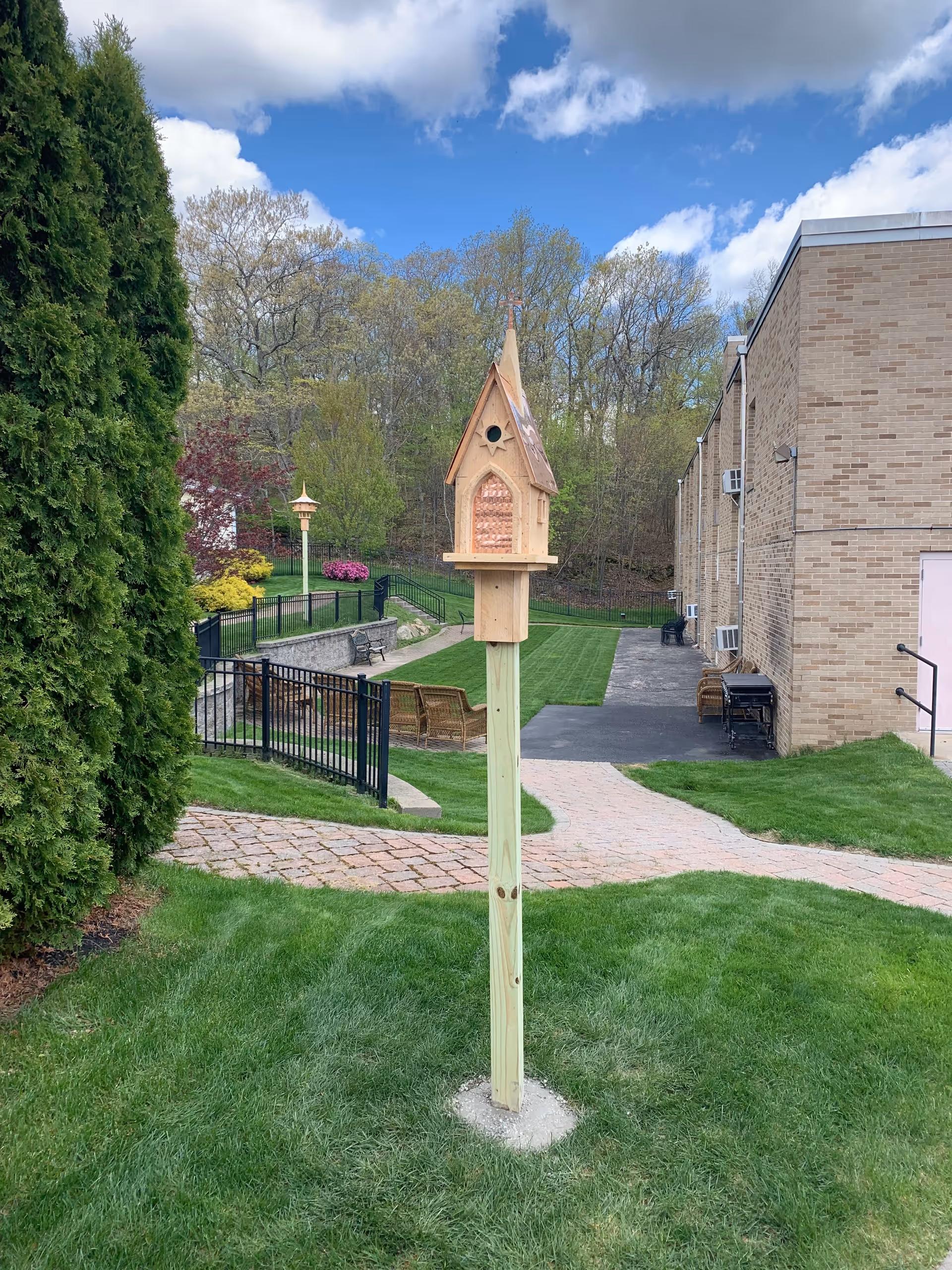 A small wooden birdhouse mounted on a tall wooden post in a grassy area outside. In the background, there is a brick building, paved walkways, benches, a black metal fence, and trees with spring foliage under a partly cloudy blue sky.