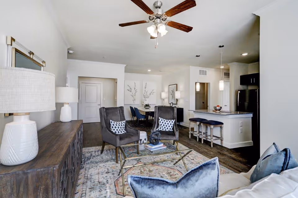 Open-concept living room with chairs, a glass coffee table on a patterned rug, ceiling fan, and a kitchen island with stools in the background.