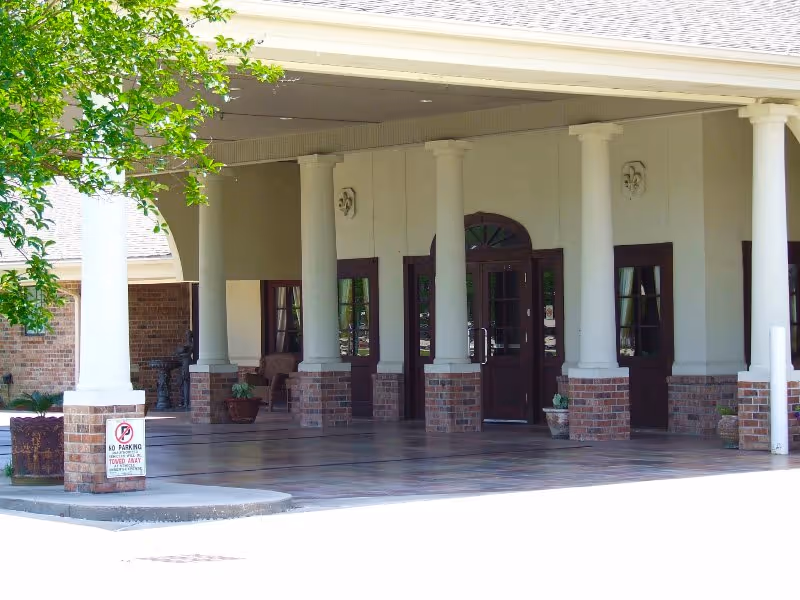 Entrance area of a building with white columns and brick bases, a covered porch with tiled flooring, several doors with glass panels, potted plants, and a 'No Parking Tow Away' sign near the curb.