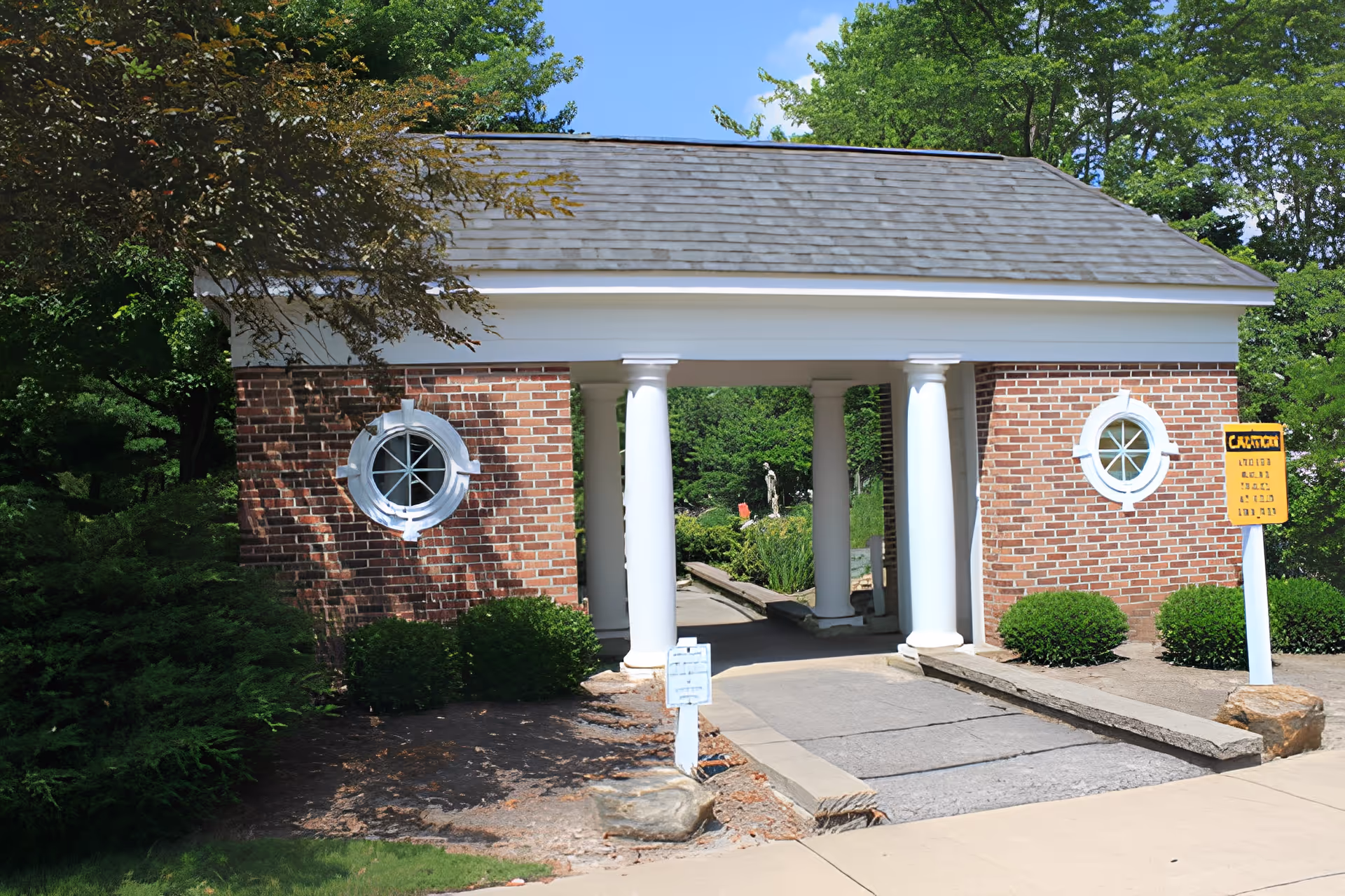 A brick structure with a shingled roof and four white columns forming a covered walkway. There are two round windows with white trim on either side of the walkway. Green bushes and trees surround the structure, and a paved path leads through it. A yellow caution sign is visible on the right side near the walkway.