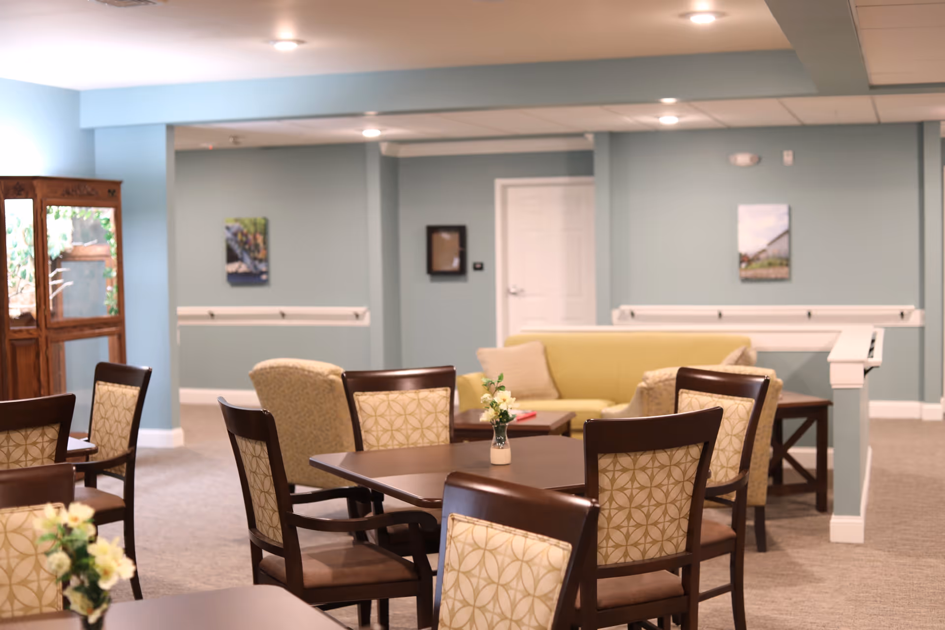 A cozy common area in a senior living facility with several wooden tables and cushioned chairs arranged around them. In the background, there is a yellow sofa with two beige armchairs and a small coffee table. The walls are painted light blue with framed pictures hanging, and there is a wooden cabinet on the left side near a window.