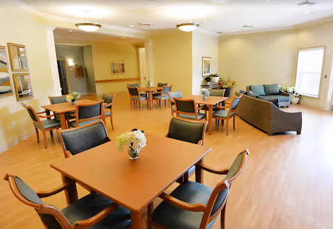 A bright and spacious common area with several wooden tables and chairs arranged for seating. Each table has a small floral centerpiece. In the background, there are two sofas and a large window letting in natural light. The room has light-colored walls and wooden flooring, creating a warm and welcoming atmosphere.