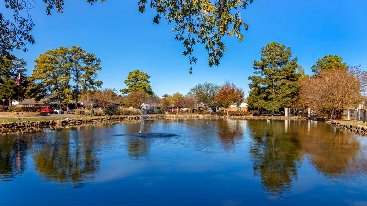 A calm pond with a small fountain, surrounded by trees and low buildings under a clear blue sky.