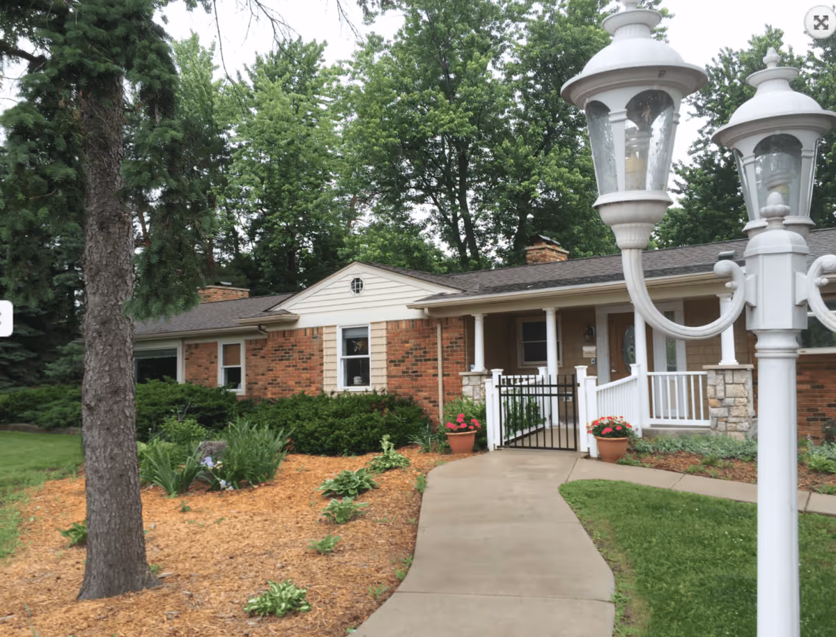 Exterior view of Silverbell Manor, a single-story brick building with a front porch, white railing, and a concrete walkway leading to the entrance. The surrounding area features green grass, trees, shrubs, and flower pots with red flowers. A white street lamp with two lanterns is visible in the foreground.