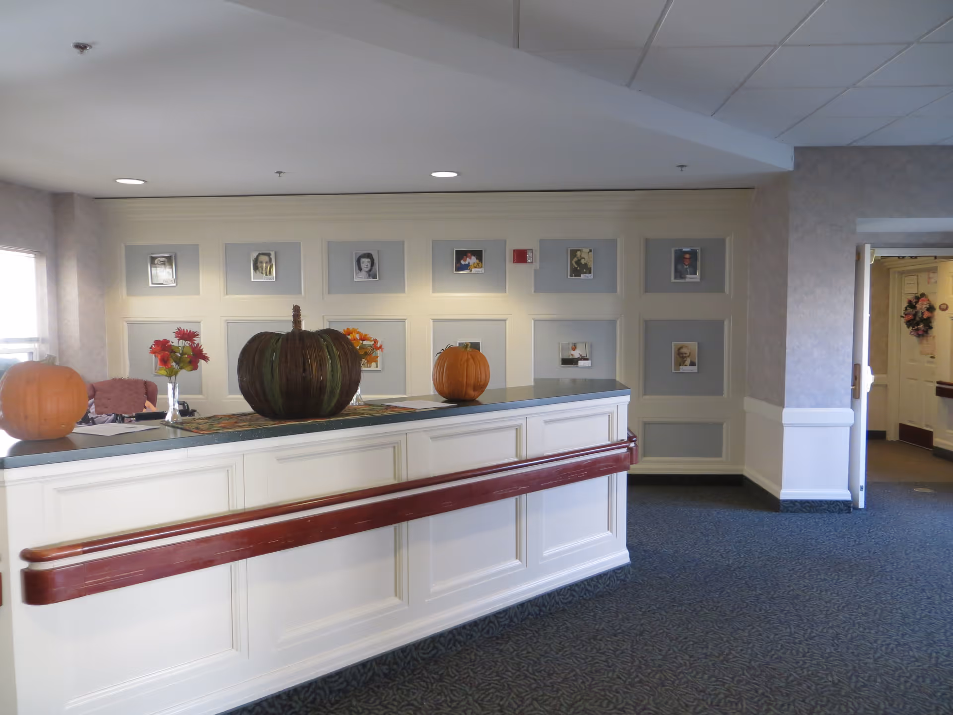 Reception desk in a senior living facility lobby decorated with pumpkins and framed photos on the wall.