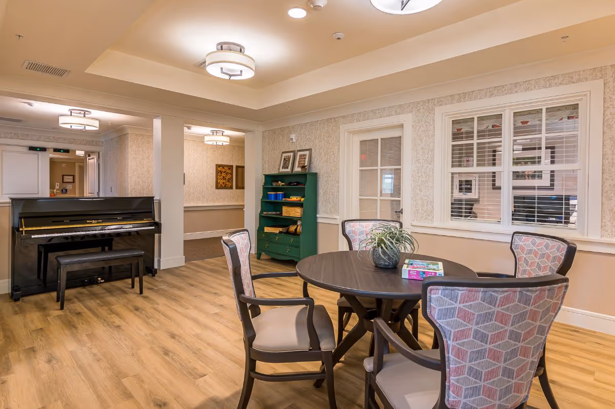 A cozy interior room with a round wooden table surrounded by four cushioned chairs with patterned upholstery. On the table, there is a small plant and a box of puzzles or games. To the left, there is a black upright piano with a matching bench. The room has light-colored walls with a chair rail, wood flooring, and ceiling lights. A green shelving unit with various items is against the far wall, and there is a window with white blinds and a door next to it.