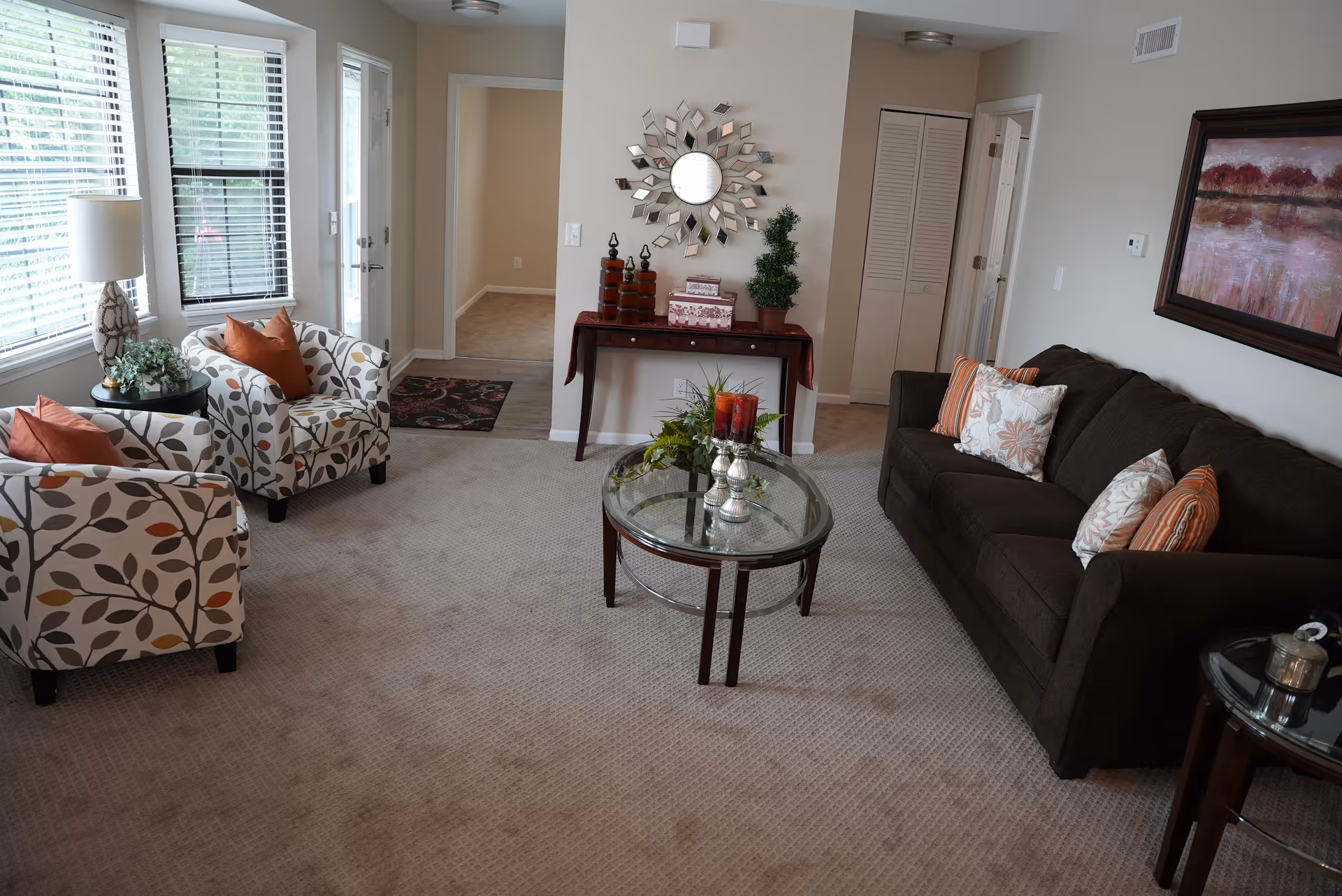 A cozy living room with a dark brown sofa adorned with patterned and striped pillows, two armchairs with leaf patterns and orange cushions, a glass coffee table with decorative candles and greenery, a side table with a lamp and plant, a console table with decorative items and a sunburst mirror above it, beige carpet, and a painting on the wall. Large windows with blinds allow natural light to fill the room.