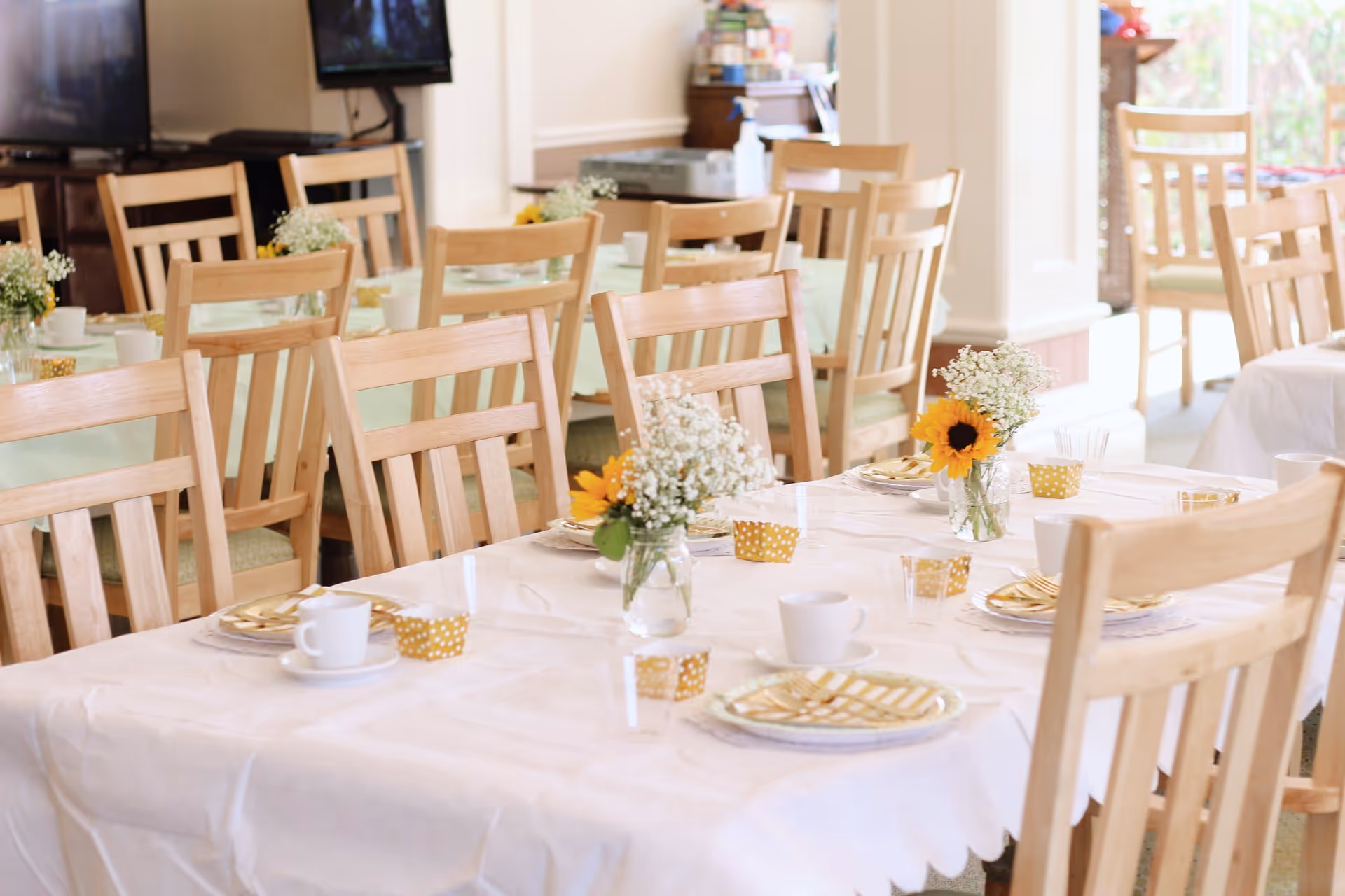 A dining room set up with multiple wooden chairs around tables covered with white and light green tablecloths. The tables are decorated with small vases of flowers including sunflowers and baby's breath, along with plates, cups, and gold-striped napkins. The room is bright with natural light coming through windows.