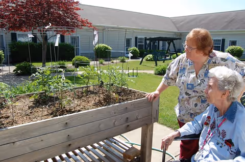 Two elderly women outdoors near a raised garden bed with plants growing. One woman is standing and the other is seated in a wheelchair. They are in a courtyard area with a building and a swing set in the background under a clear sky.