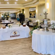 Interior view of a senior living facility lobby or common area with tables covered in white cloths, one displaying a banner with the Summerset logo and name. The space is decorated with flowers, plants, and a decorative water fountain, with seating and lighting visible in the background.