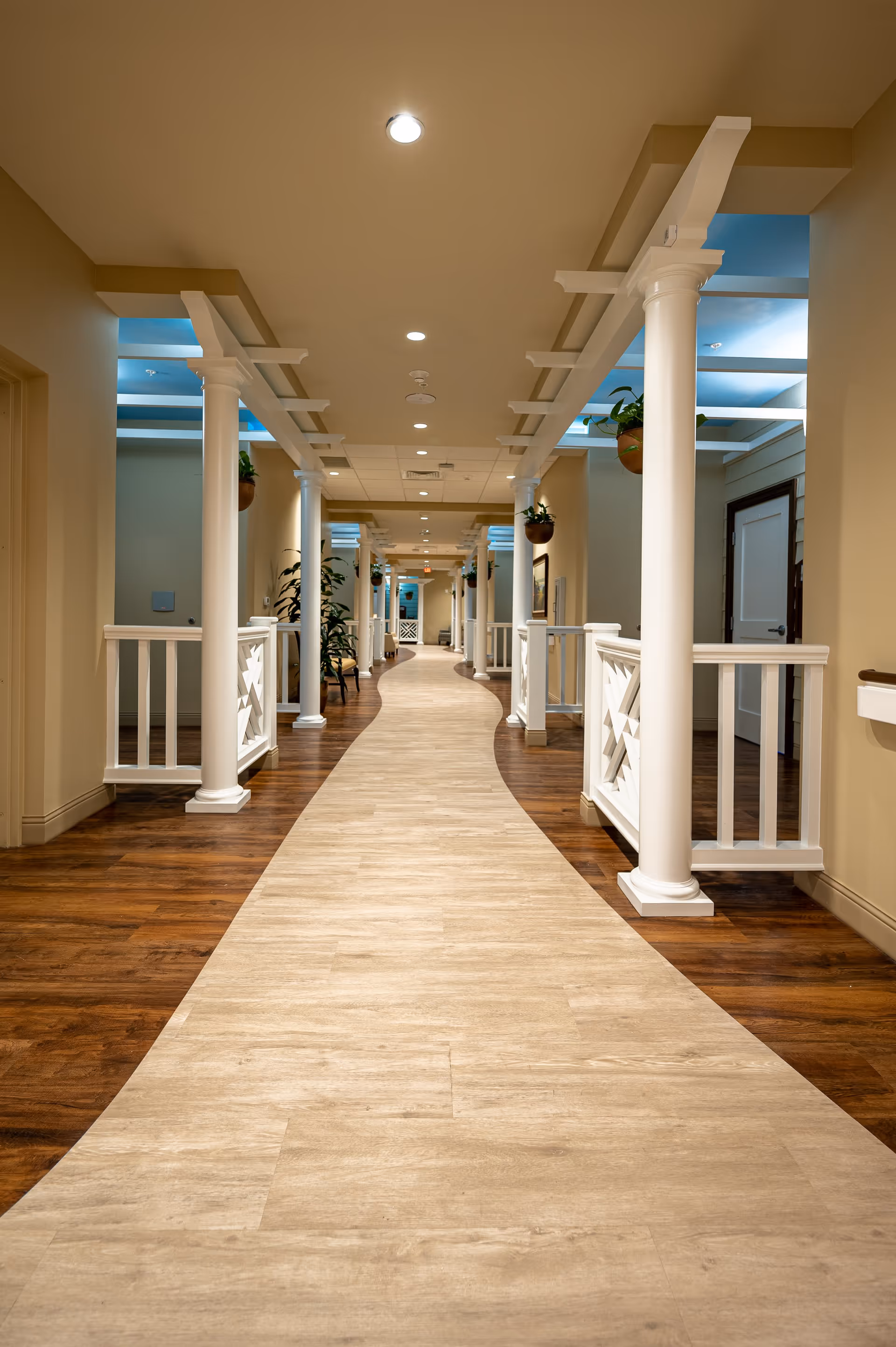 A well-lit indoor hallway with a curved light wood floor path flanked by darker wood flooring. The hallway features white columns and railings, with potted plants hanging from the ceiling and placed along the sides. The ceiling has recessed lighting and a beige color scheme.