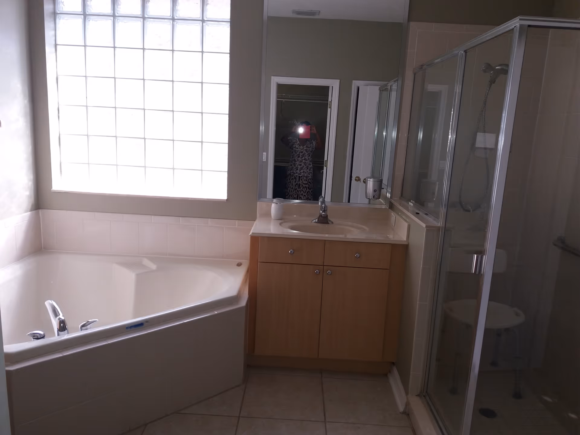 Bathroom with a large corner bathtub under a frosted glass window, a wooden vanity with a sink and faucet, a mirror above the sink, and a glass-enclosed shower with a showerhead and a white stool inside.