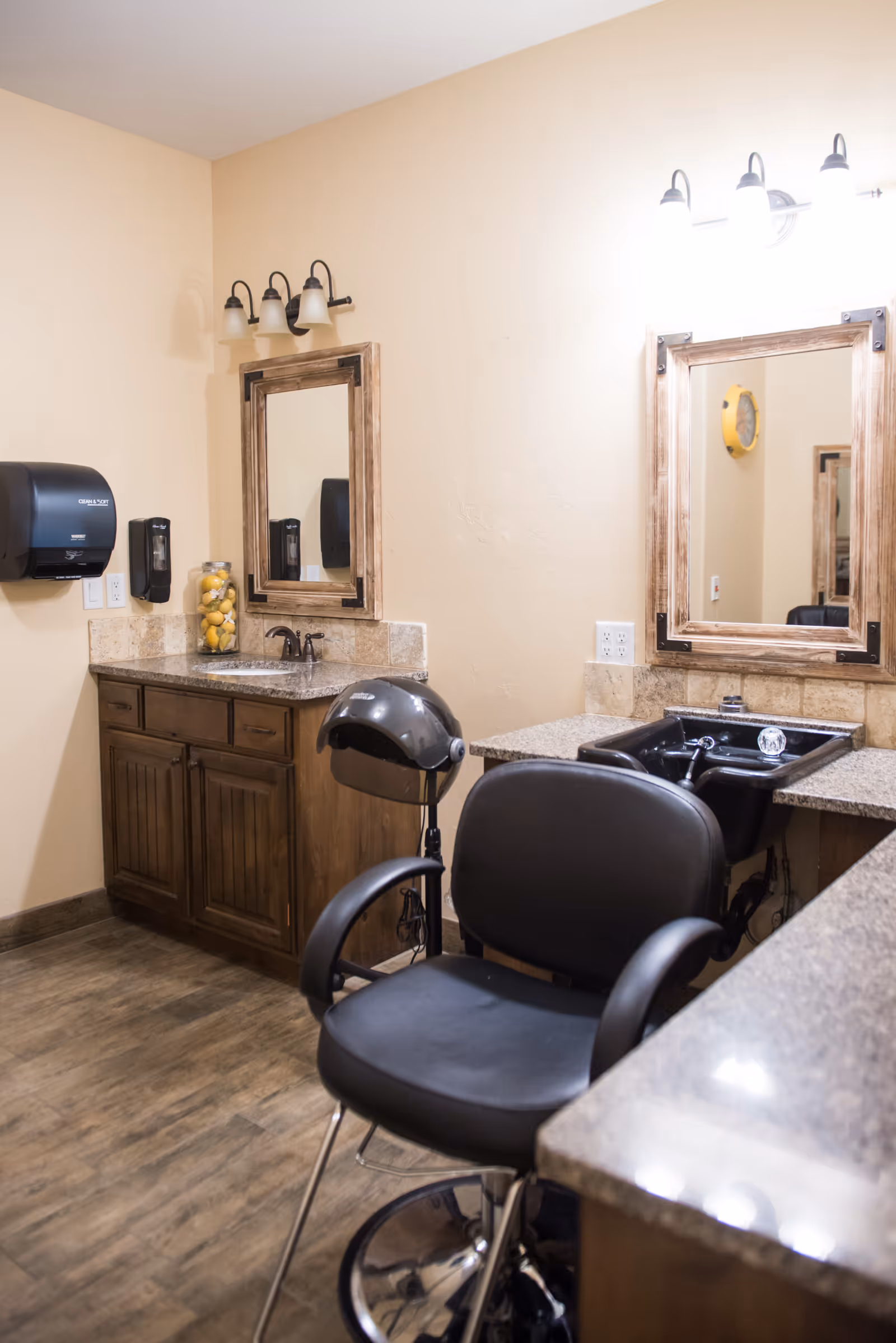 Interior view of a salon area in an assisted living facility featuring a black salon chair, a hair dryer, two wooden framed mirrors above granite countertops with sinks, wooden cabinets below, and wall-mounted soap and paper towel dispensers.