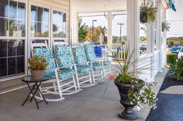 A covered porch area with four white rocking chairs featuring blue and green floral cushions. There is a small round table with a potted plant on it next to the chairs. The porch is decorated with hanging plants and large potted plants, and there is a view of a parking lot and trees in the background.