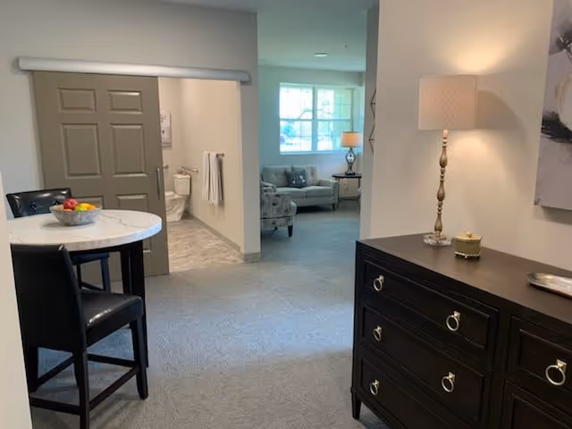 Interior view of a senior living suite with a small round dining table and chairs, a dark dresser with a lamp, and a seating area visible down a short hallway.