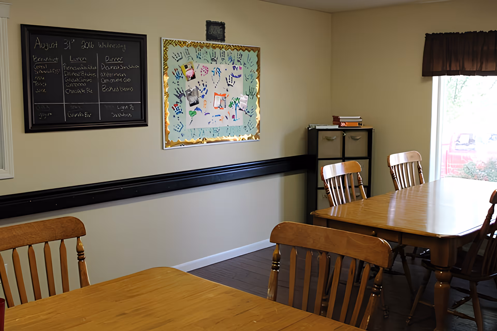 A dining room with wooden tables and chairs. On the wall, there is a blackboard with a handwritten menu dated August 31, 2016, and a bulletin board decorated with colorful handprints and photos. A window with a dark valance lets in natural light, and a small storage unit with fabric drawers is in the corner.