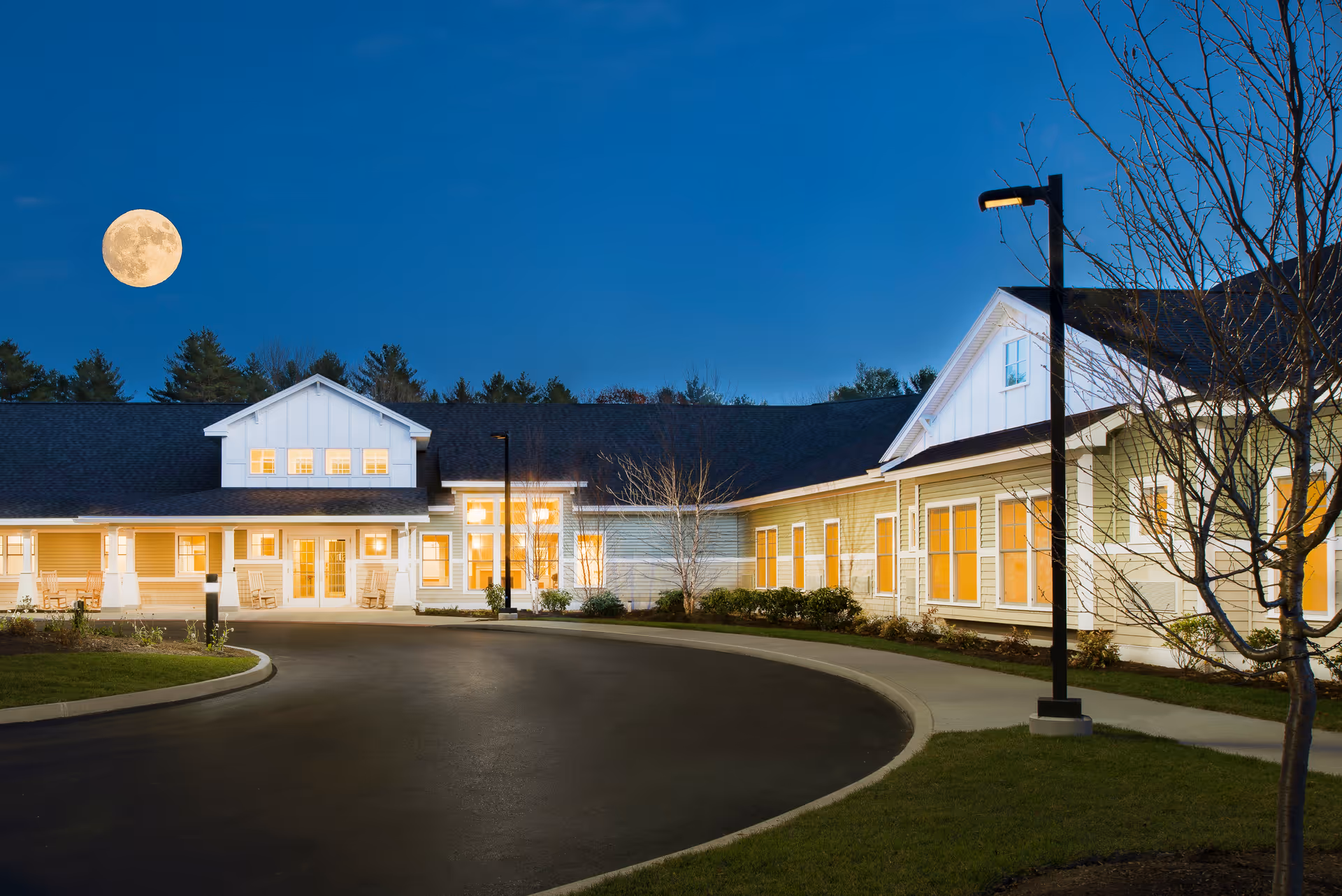 Exterior view of Avita of Brunswick senior living facility at dusk with lights glowing from windows, a curved driveway, street lamps, and a full moon in the clear sky.