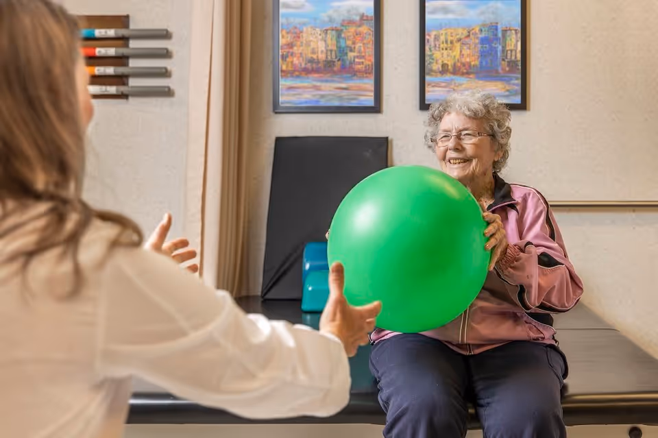 An elderly woman wearing glasses and a pink jacket is sitting on a padded bench holding a large green exercise ball, smiling as she interacts with another person whose back is to the camera. The setting appears to be a rehabilitation or therapy room with colorful paintings on the wall and exercise equipment nearby.